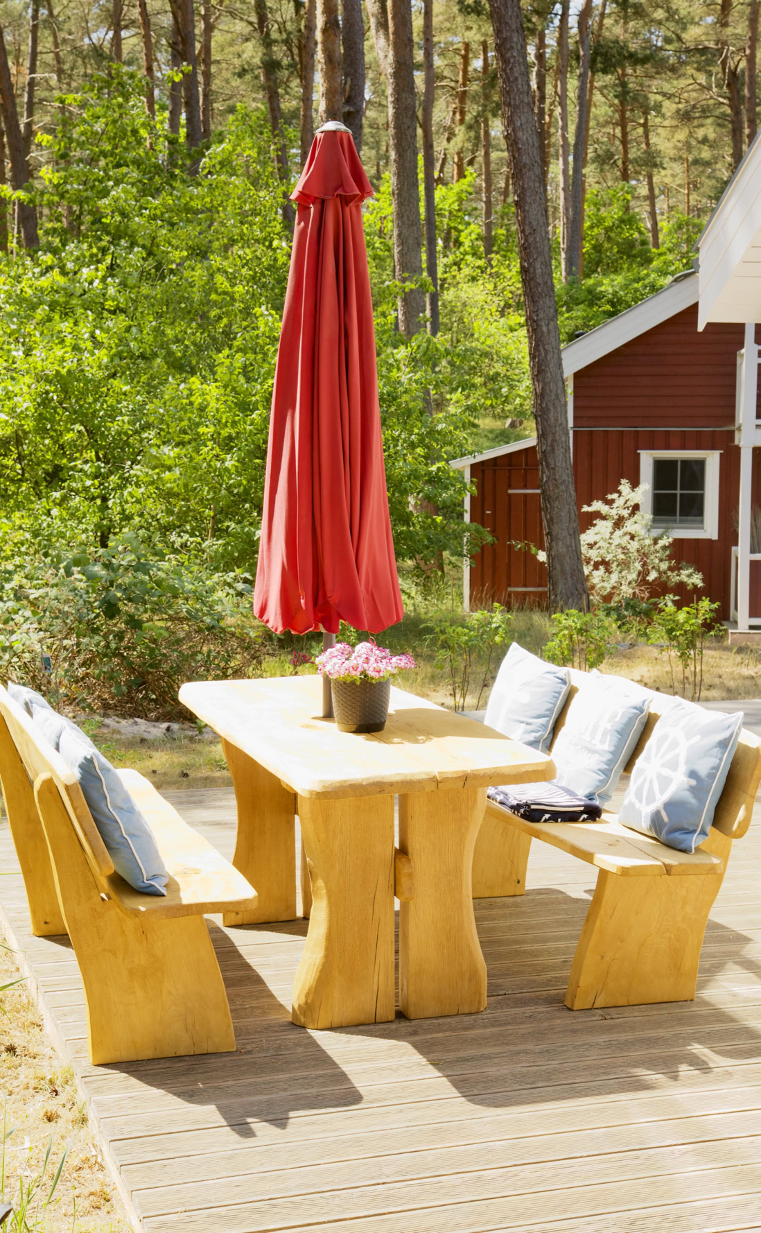 Holztisch mit Bänken und Kissen auf einer Terrasse, roter Sonnenschirm, Bäume und rotes Gebäude im Hintergrund.