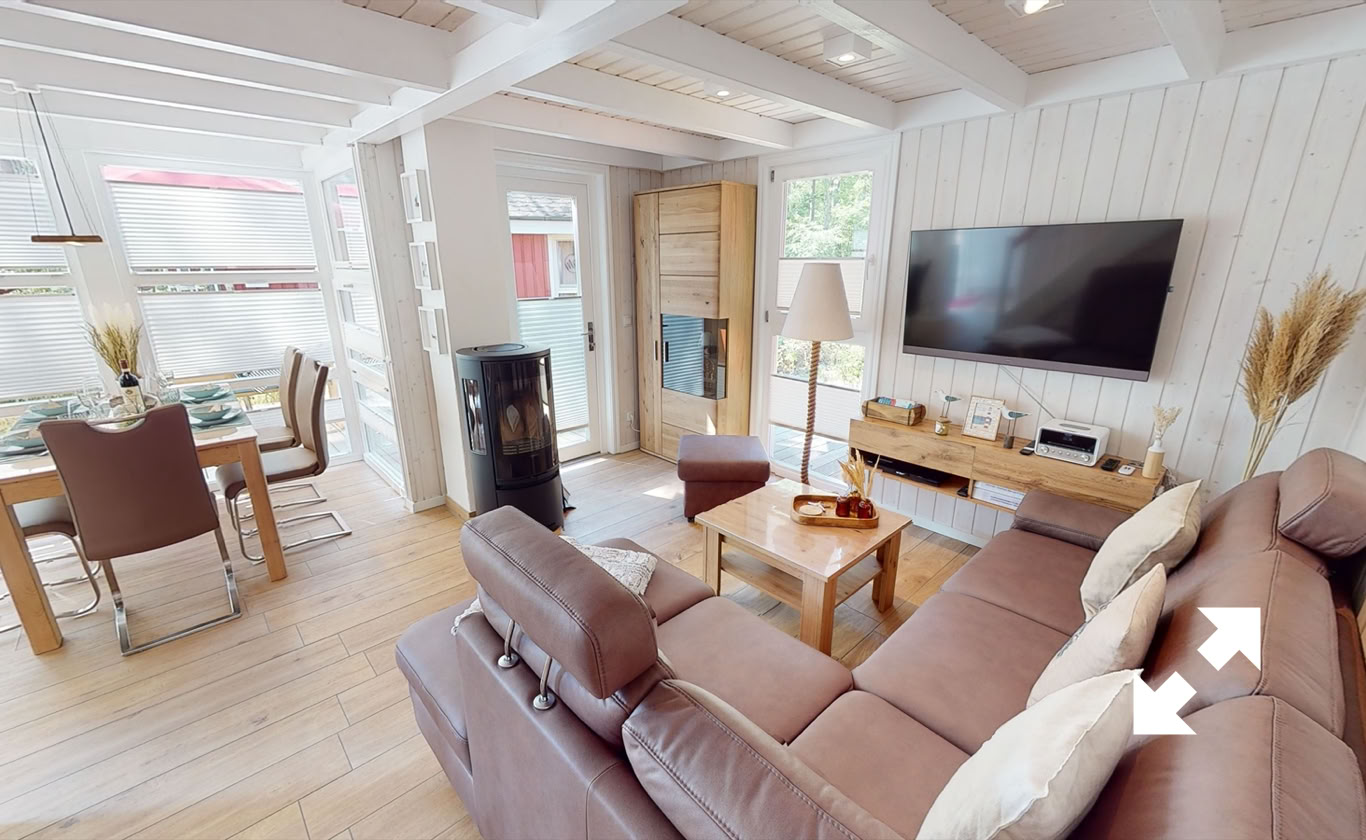 Wohnzimmer mit braunem Sofa, Holztisch, großem Fernseher an der Wand, Kaminofen, Esstisch mit Stühlen und großen Fenstern.
