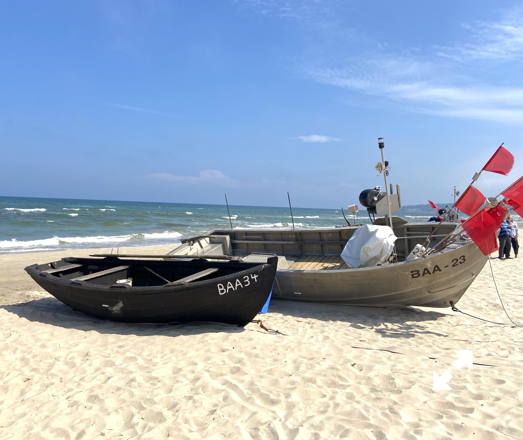 Zwei Boote am Sandstrand, eines schwarz und eines grau, mit roten Fahnen, im Hintergrund das Meer und blauer Himmel.