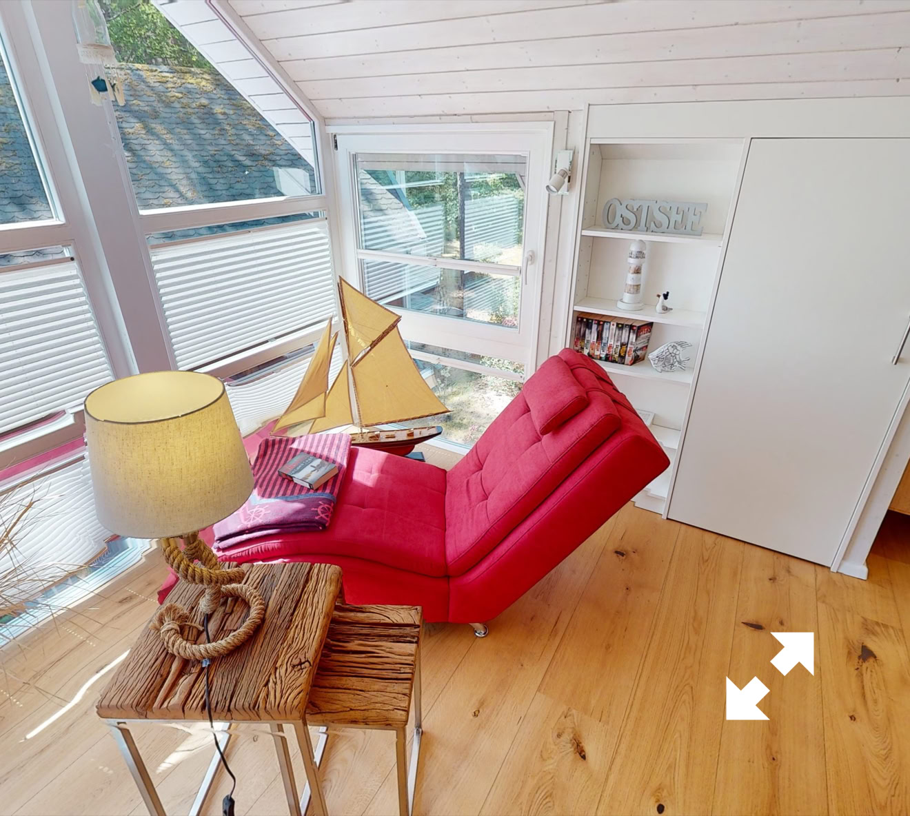 Gemütlicher Raum mit einem roten Sessel, einem kleinen Tisch mit Lampe, einem Modellsegelboot und großen Fenstern. Heller Holzboden und weiße Wände.