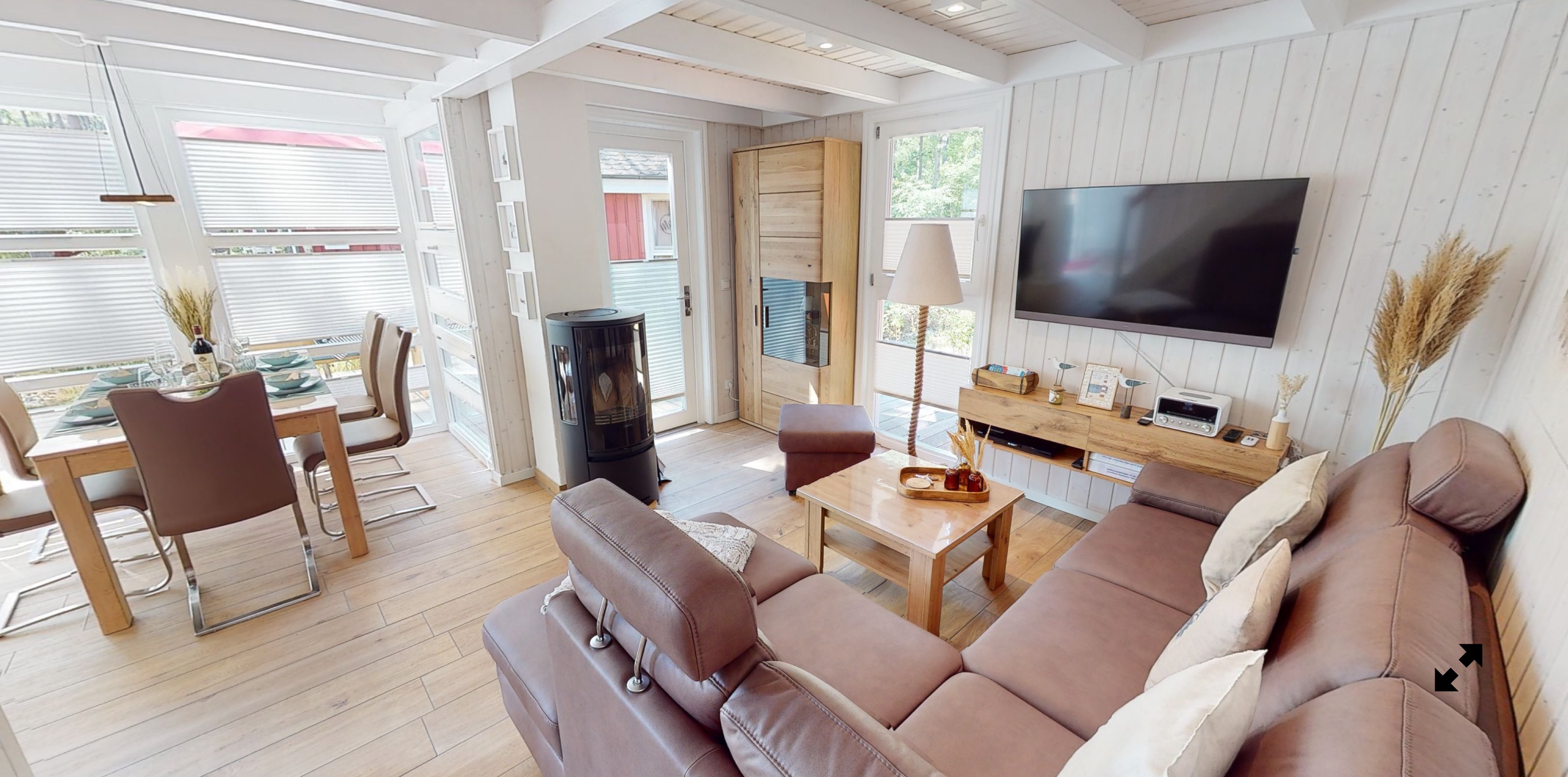 Wohnzimmer mit braunem Sofa, Holztisch, großem Fernseher an der Wand, Kaminofen, Esstisch mit Stühlen im Hintergrund und Dekorationen auf dem Tisch und Regal.