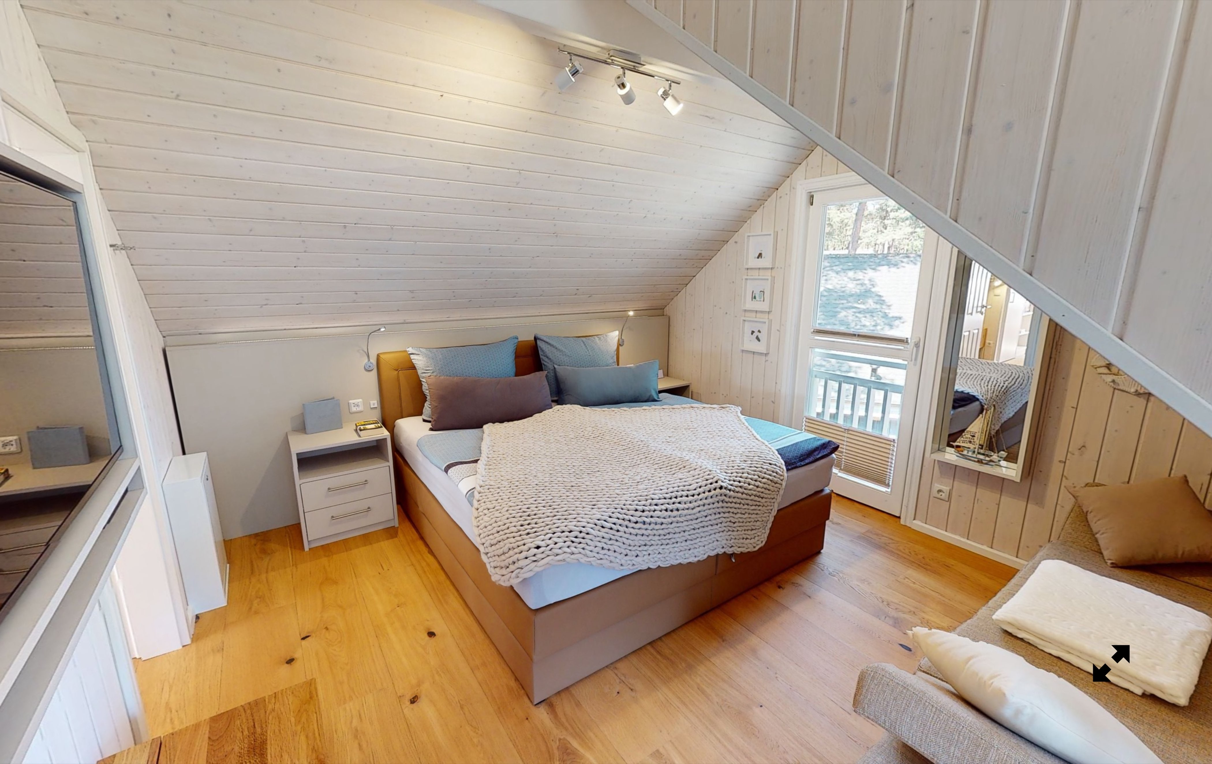 Gemütliches Schlafzimmer mit Holzboden, einem Doppelbett mit blauen und weißen Kissen, zwei Nachttischen, einem großen Spiegel und einem Fenster mit Blick nach draußen.