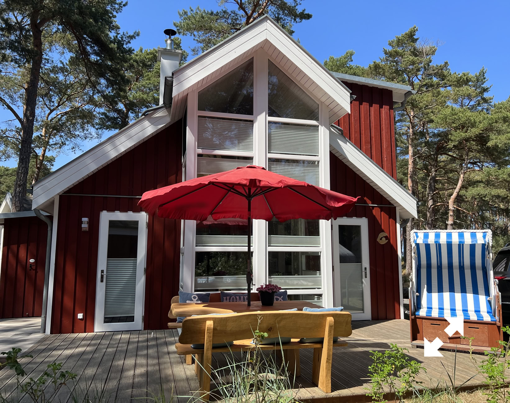 Rotes Holzhaus mit großen Fenstern, rotem Sonnenschirm, Holztisch und Strandkorb im Vordergrund, umgeben von Bäumen.