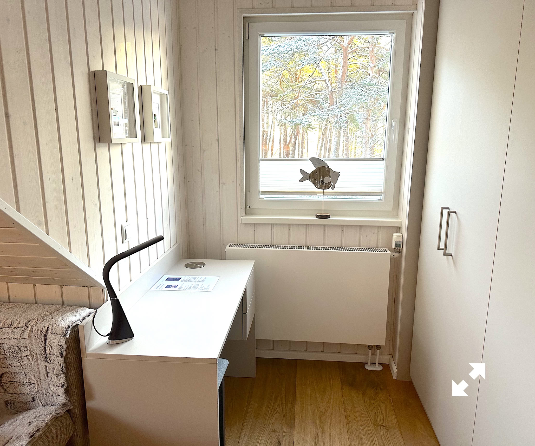 Kleines Zimmer mit einem weißen Schreibtisch, einer schwarzen Lampe, einem Sofa und einem Fenster mit Blick nach draußen.