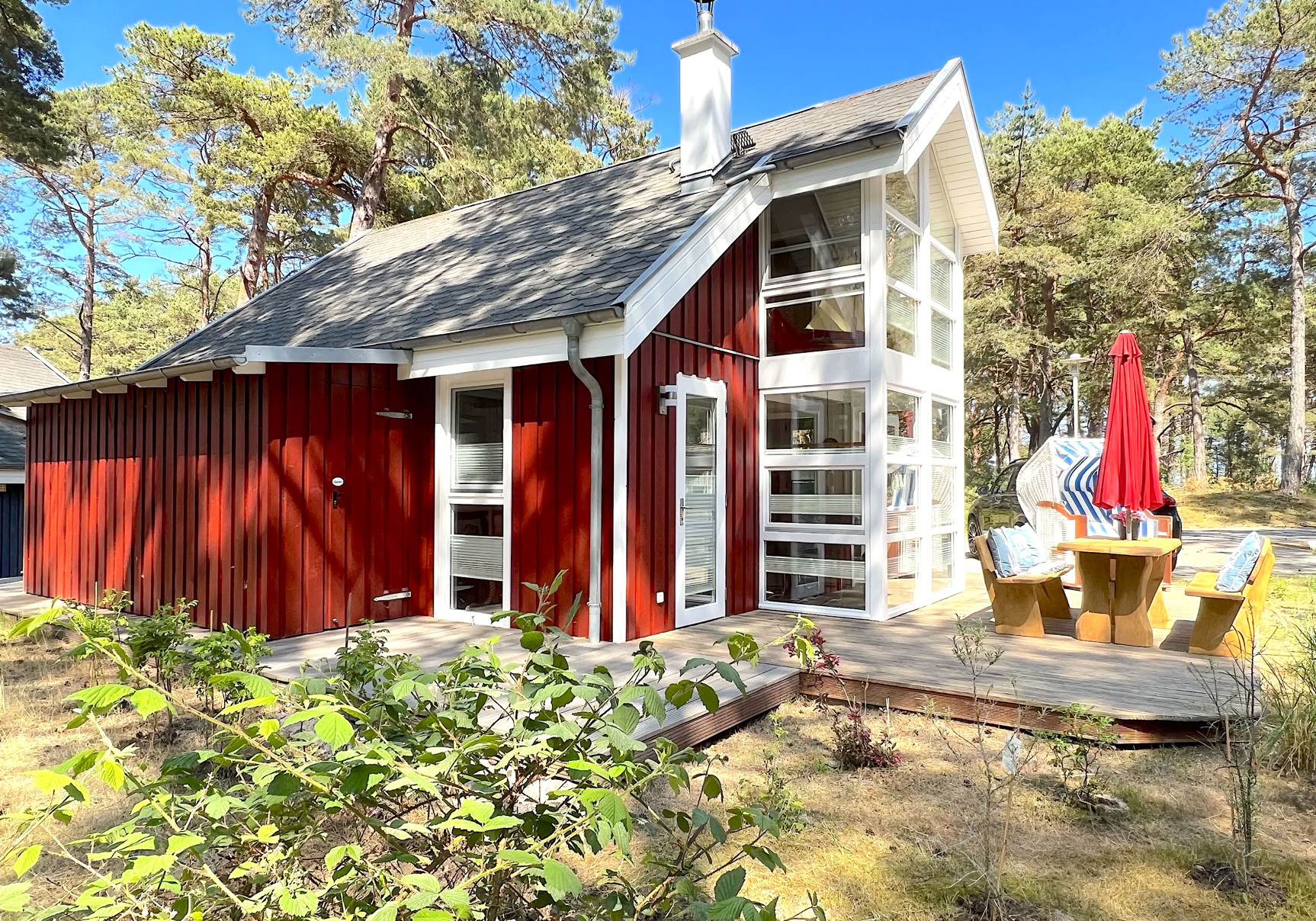 Rotes Holzhaus mit großen Fenstern, umgeben von Bäumen, mit einem Tisch und Stühlen im Freien.