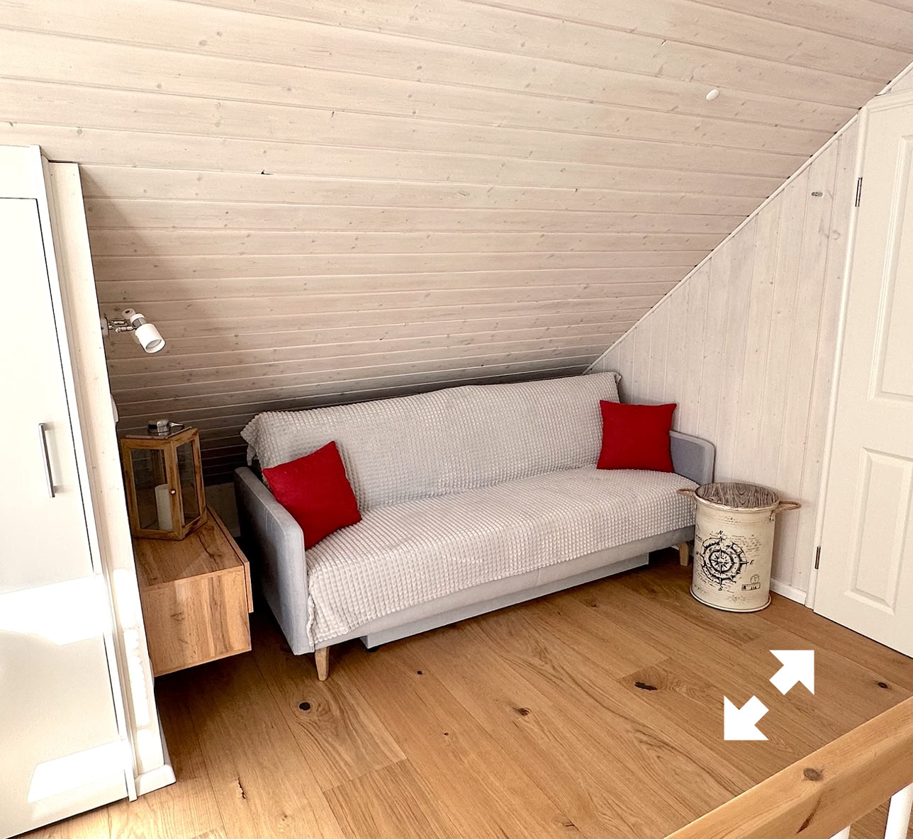 Ein Raum mit Holzdecke und Holzboden, einem weißen Sofa mit roten Kissen, einem kleinen Holztisch, einem weißen Schrank und einem dekorativen Korb.