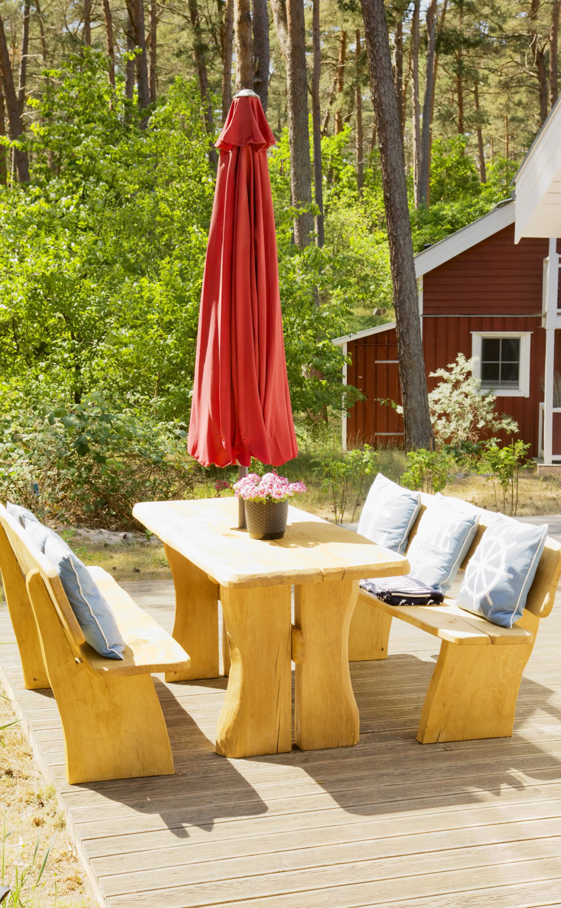 Holztisch mit Bänken und Kissen auf einer Terrasse, roter Sonnenschirm, Bäume und rotes Gebäude im Hintergrund.