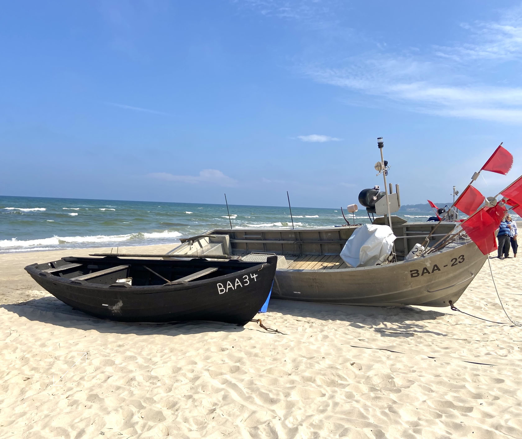 Zwei Boote am Sandstrand, eines schwarz und eines grau, mit roten Fahnen, im Hintergrund das Meer und blauer Himmel.